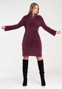 Strukturiertes, übergroßes, lila gestricktes Kleid mit hohem Kragen und langen Ärmeln, ergänzt durch kniehohe schwarze Stiefel.