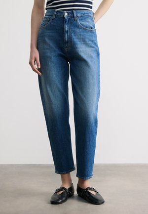 Άτομο που φοράει μπλε τζιν με ψηλή μέση και ίσια μπατζάκια, μαύρα επίπεδα παπούτσια με λουράκια και ριγέ τοπ, στέκεται σε γκρίζο πάτωμα.