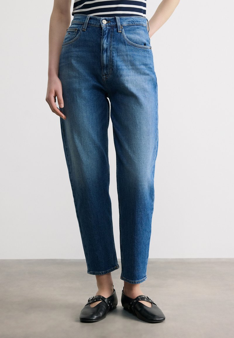 Άτομο που φοράει μπλε τζιν με ψηλή μέση και ίσια μπατζάκια, μαύρα επίπεδα παπούτσια με λουράκια και ριγέ τοπ, στέκεται σε γκρίζο πάτωμα.