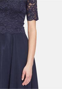 Námornícky modré šaty s čipkovaným korzetom zdobeným kvetinovými vzormi a krátkymi rukávmi, splývavá sukňa z hladkej tkaniny a priliehavý pás.