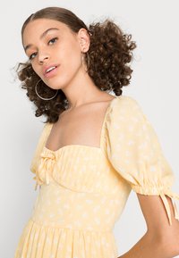 Vestido amarelo com flores, mangas balão, punhos elásticos, decote quadrado e detalhe de atar à frente; tecido leve com padrão branco subtil.