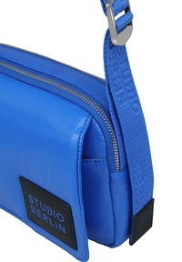 Borsa crossbody blu con finitura lucida, chiusura con zip, tracolla regolabile e una patch nera con il marchio "STUDIO BERLIN."