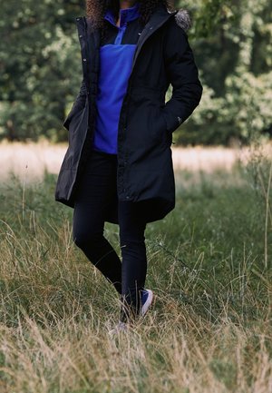 Persona con una parka negra con capucha con borde de piel y un suéter azul, de pie con las manos en los bolsillos en hierba alta al aire libre.