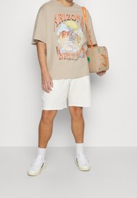 Beige graphic t-shirt with a desert illustration, white shorts, and white sneakers. Includes a beige bag with orange accents and strap.