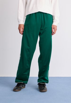 Grønne, løse joggebukser laget av glatt stoff, med snor i livet og stripesider, kombinert med svarte sko.