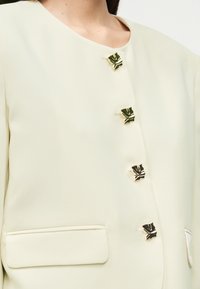 Φωτεινό πράσινο σακάκι με απαλή υφή, που διαθέτει πέντε κλεισίματα με χρυσές λεπτομέρειες και μία τσέπη στην αριστερή πλευρά. Στρογγυλεμένος σχεδιασμός λαιμού.