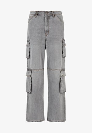 Mężczyzna w jasnoszarej oversize koszulce z napisem „89 Studios” i luźnych szarych spodniach typu cargo oraz białych sneakersach, stojący z jedną ręką w kieszeni.