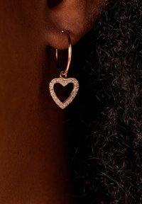 Ohrringe aus Roségold mit einem herzförmigen Anhänger, der mit klaren Kristallen verziert ist und ein zartes offenes Design sowie eine glänzende Textur präsentiert.
