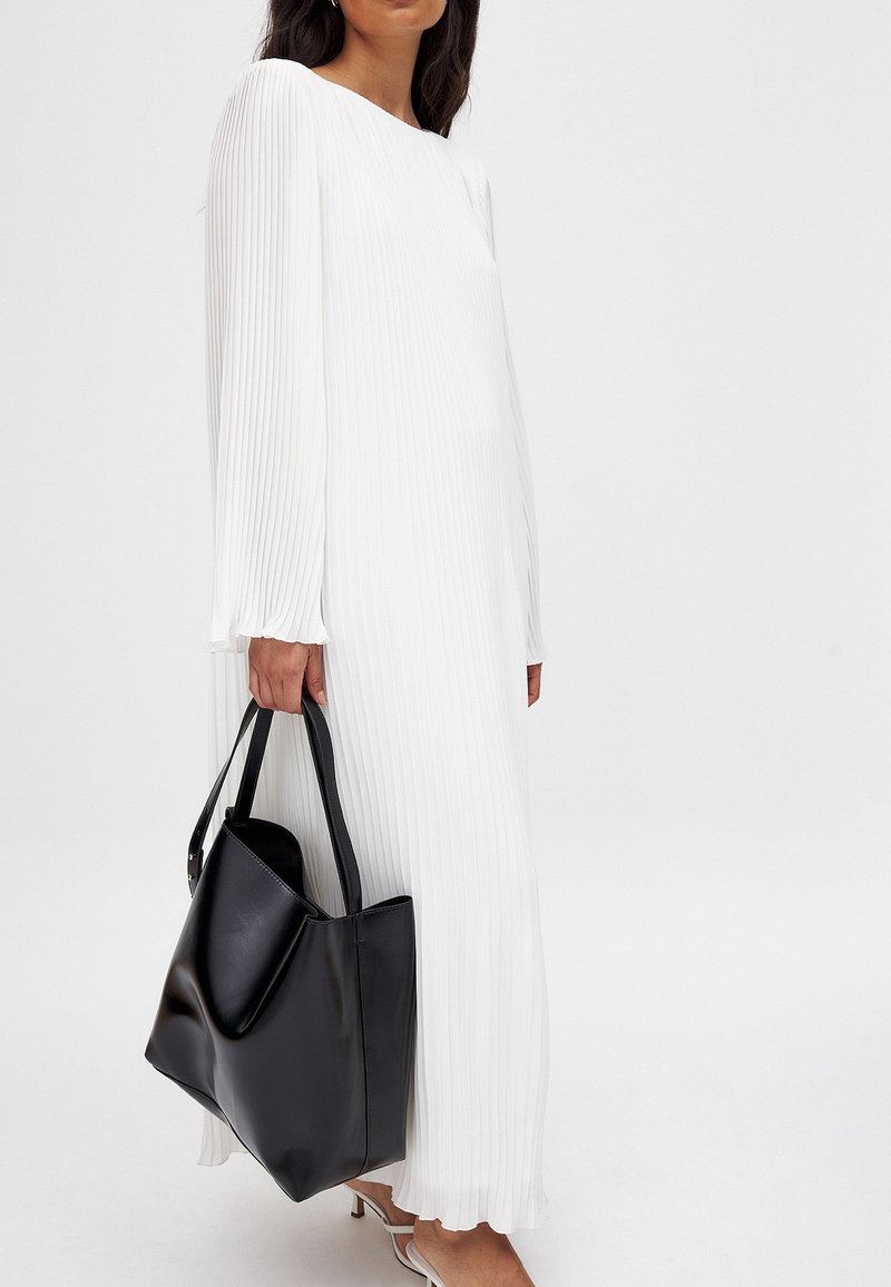 Valkoinen laskostettu maksimekko pitkillä hihoilla, yhdistetty mustan nahkakassin kanssa. Silkkinen kangas, jossa on virtaava muotoilu. Avoin kaula-aukko, minimaalinen yksityiskohtaisuus.