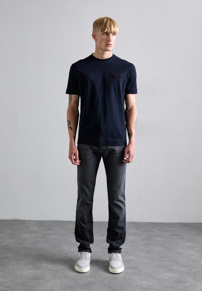 Marinblaues Kurzarm-T-Shirt mit einem strukturierten Emblem auf der Brust, kombiniert mit dunklen Jeans und grauen Sneakers. Glattes Material, taillierte Passform.