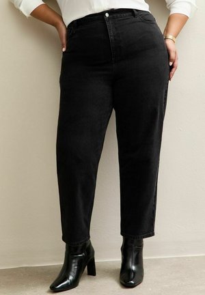 Kobieta ubrana w czarne, wysokowciskane, zwężane dżinsy, białą bluzkę oraz czarne botki na obcasie, stojąca z rękami na biodrach.