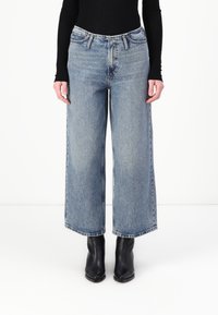 Videbeinede lyseblå jeans laget av denim, med høy midje, flere lommer og rå sømfinish, sammen med svarte ankelstøvler.