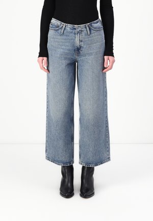 Videbeinede lyseblå jeans laget av denim, med høy midje, flere lommer og rå sømfinish, sammen med svarte ankelstøvler.