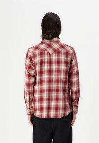 Rot-beige kariertes Flanellhemd mit spitzem Schulterbereich, geknöpften Manschetten und abgerundetem Saum, von hinten präsentiert.