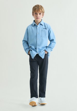 Niño de pie de frente con una camisa azul claro abotonada, pantalones negros y zapatillas blancas, con las manos en los bolsillos sobre un fondo liso.