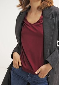 Grau Wollmantel über einem burgunderfarbenen Satinoberteil mit entspanntem Ausschnitt. Dunkelblaue Jeans mit leichtem Stretch. Minimalistisches Design, glatte Textur.