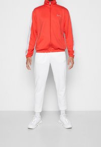 Chaqueta de chándal roja con franjas laterales blancas, cremallera completa, cuello alto, combinada con pantalones blancos ajustados y zapatillas deportivas blancas.