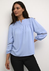 Blusa azzurra chiara realizzata in tessuto morbido, con pieghe sul petto, colletto rotondo e dettagli in pizzo sulle maniche. Design dal taglio morbido.