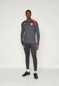 Mies, joka on pukeutunut tummanharmaaseen ja mustaan urheiluharjoitusasuun, jossa on punaisia yksityiskohtia ja urheilulogoja, seisoo vaaleaa yksinkertaista taustaa vasten.