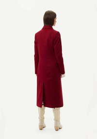 Röd ullkappa med en lång silhuett, ryggslits och avskuren krage. Modell som bär beigefärgade knälånga stövlar. Enkel vit bakgrund.