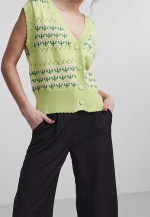 Person iført lys grøn ærmeløs strikvest med blomstermønster og store knapper, kombineret med løstsiddende sorte bukser og hænderne i lommerne.