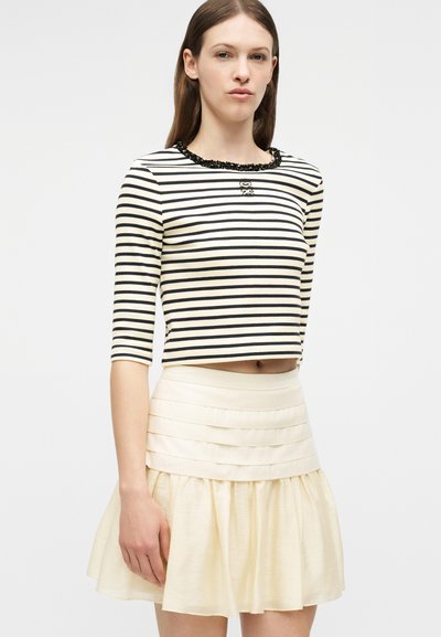Γυναίκα που φοράει μια ριγωτή μαύρη και λευκή crop top με μανίκια μέχρι τον αγκώνα και μια κρεμ μίνι φούστα με βολάν, σε απλό φόντο.