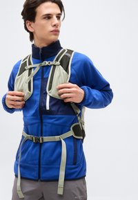 Νεαρός άνδρας που ρυθμίζει τους ιμάντες μιας ανοιχτόχρωμης γκρι σακίδας, φορώντας μπλε φλις μπουφάν και γκρι παντελόνι, στέκεται μπροστά σε λευκό απλό φόντο.