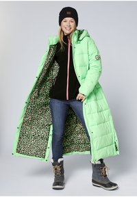 Langer, mintgrüner Steppmantel mit einem Futter in Leopardenmuster, schwarzem Reißverschluss-Top, blauen Jeans und grauen Winterstiefeln mit weißem Pelzbesatz.