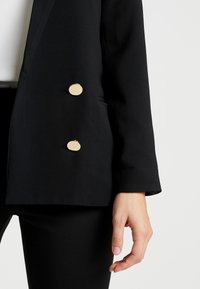 Černý blazer s přizpůsobeným střihem, dvouřadým zapínáním se dvěma kulatými zlatými knoflíky a hladkou texturou materiálu.