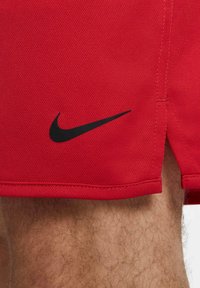Primo piano di una gamba pelosa di una persona che indossa pantaloni corti rossi Nike con il logo Swoosh nero vicino all'orlo.