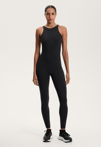 Fekete, testhez simuló bodysuit ujjatlan kialakítással, magas nyakkal és varrat nélküli textúrával, fekete sportcipőkkel párosítva.