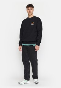 Zwarte sweatshirt met een graphic logo op de borst, gecombineerd met donkere spijkerbroeken en zilveren sportschoenen. Model staat tegen een witte achtergrond.