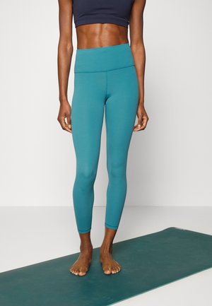 Teal, high-waisted leggings lavet af strækbart stof, tætsiddende design, glat tekstur, sammen med en mørk cropped top, barfodet på en yogamåtte.