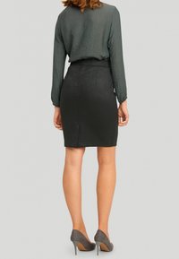 Černá tužková sukně s hladkým povrchem, vysokým pasem a rozparkem vzadu, spárovaná s tmavozelenou blůzou s dlouhým rukávem a šedými vysokými podpatky.
