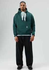 Donkergroene hoodie met voorzak en een witte bandana-stijl accent. Gecombineerd met losse zwarte broek en zwarte robuuste schoenen.