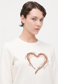 Kremgenser med et paljett hjerte-design i roségull. Glatt tekstur, rund hals, og lange ermer.