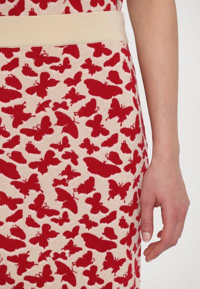 Rode vlinderprint op beige stof, met een zachte textuur en een brede beige tailleband. Close-up van een hand dichtbij de stof.
