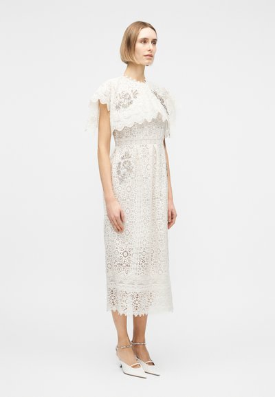 Γυναίκα που φοράει λευκό δαντελένιο φόρεμα μήκους μέχρι τη μέση της γάμπας με κοντά μανίκια και ανθικά σχέδια, συνδυασμένο με λευκά μυτερά τακούνια με λουράκια στον αστράγαλο.