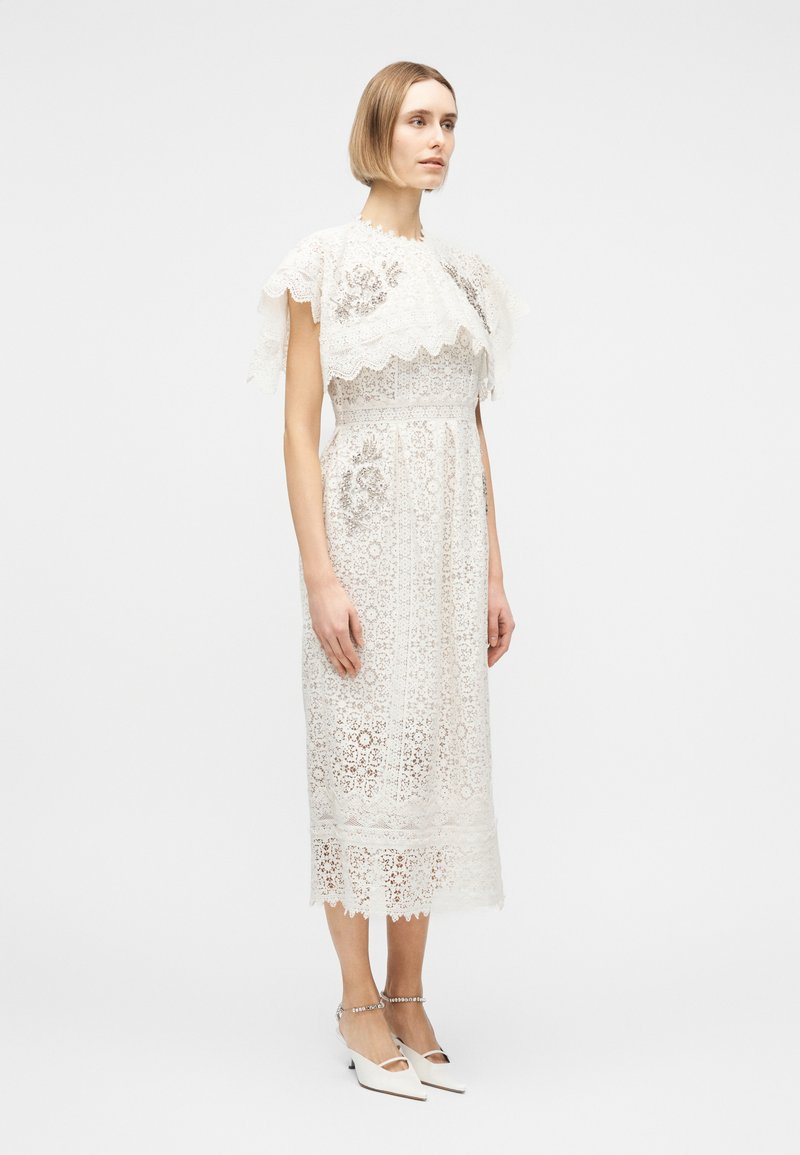 Frau trägt ein weißes Midikleid aus Spitze mit kurzen Ärmeln und Blumenmustern, kombiniert mit weißen spitz zulaufenden Schuhen mit Knöchelriemen.