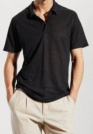 Schwarzes Kurzarm-Polohemd mit einem knöpfbaren Kragen, aus einem weichen Stoff gefertigt. Getragen mit hellen, gestreiften Hose.
