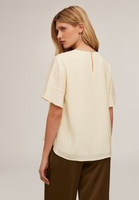 Kurzärmlige Bluse aus leichtem gelben Stoff mit einem runden Ausschnitt und einem kleinen Schlitze hinten, in lockerer Passform, kombiniert mit braunen Hosen.