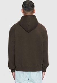 Haut à capuche de couleur marron foncé avec une grande capuche, coupe ample et poignets côtelés. Le tissu semble doux avec une texture lisse. Pas de logos ni de motifs visibles.