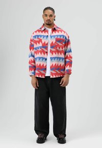 Mehrfarbiges Hemd mit Knöpfen in Rot, Blau und Weiß mit geometrischen Mustern, kombiniert mit locker sitzenden schwarzen Hosen und schwarzen Schuhen mit pinkfarbenen Schnürsenkeln.