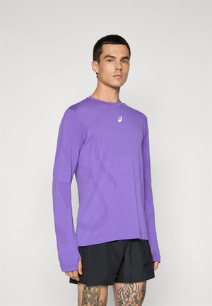 Mann, der ein lila langärmliges Sportoberteil und schwarze Shorts trägt, steht vor einem schlichten hellen Hintergrund.