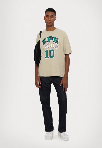 Beige graphic t-shirt featuring green and purple text reading "KPR VAULT 10," paired with dark jeans and white sneakers. Black crossbody bag on shoulder.