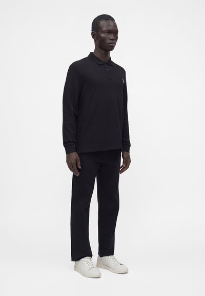 Homme debout portant un polo manches longues noir, un pantalon noir et des baskets blanches sur un fond clair uni.