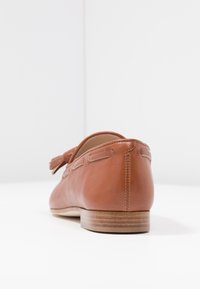 Bruine leren instapper met een gladde textuur, voorzien van een kwastje, ronde neus en een lichte houten zool. Minimale stikselaccenten.