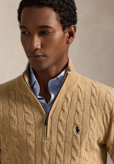 Mand iført en beige kabelstrikket lynlås-sweater over en lyseblå skjorte med knapper, der kigger til siden mod en ensfarvet baggrund.
