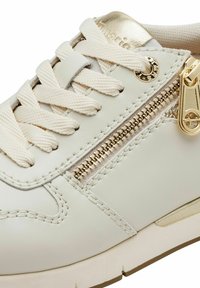 Sneaker in pelle crema con lacci crema, una cerniera laterale dorata e un accento metallico dorato sul tallone. Suola in gomma testurizzata.