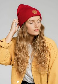 Czerwony, żeberkowany beret z okrągłą skórzaną łat�ą, noszony przez osobę z długimi, falującymi włosami. Tło jest jasnoszare. Częściowo widoczna żółta kurtka.